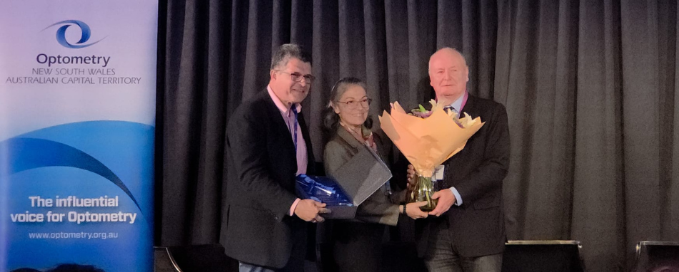 Man, woman and man standing on a stage holding an award and flowers, with black backdrop and Optometry NSW/ACT Sign in the background