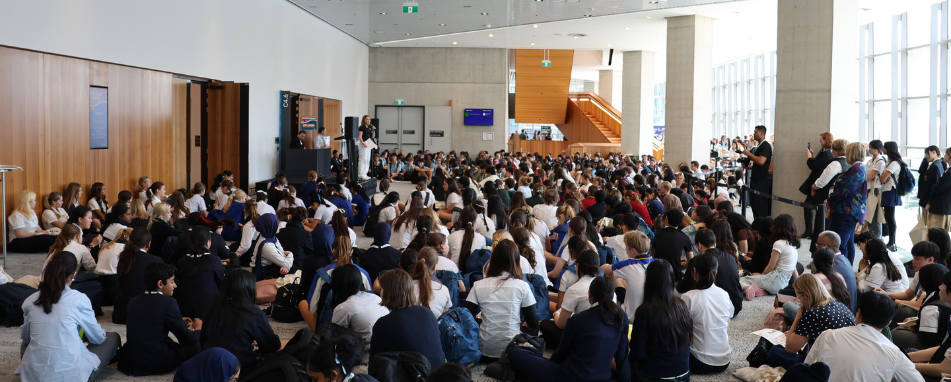 School children sitting on ground facing woman on stage speaking into microphone