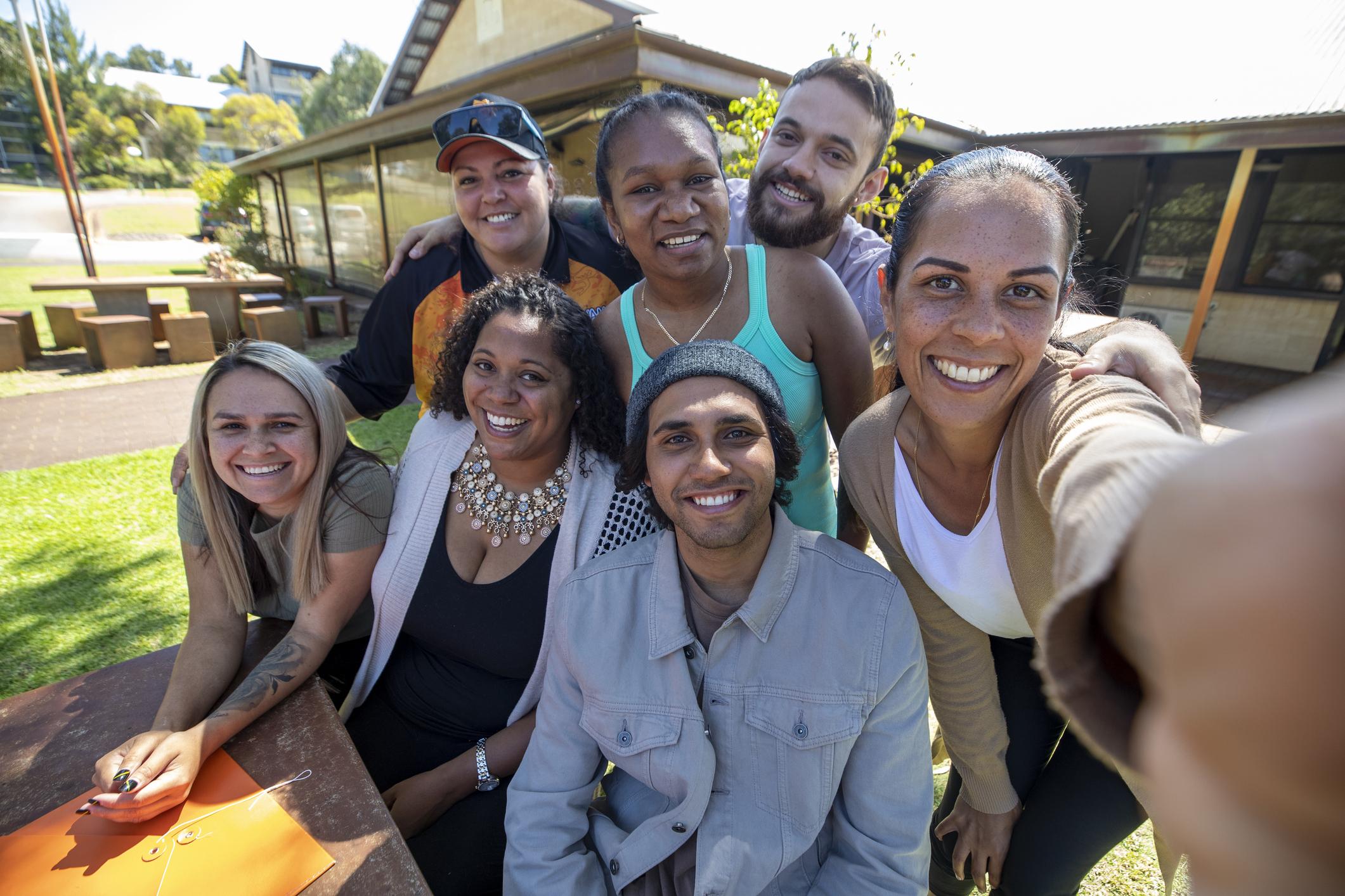 Seven people sitting and standing around a table. Person on right holding the camera to take a selfie of group.