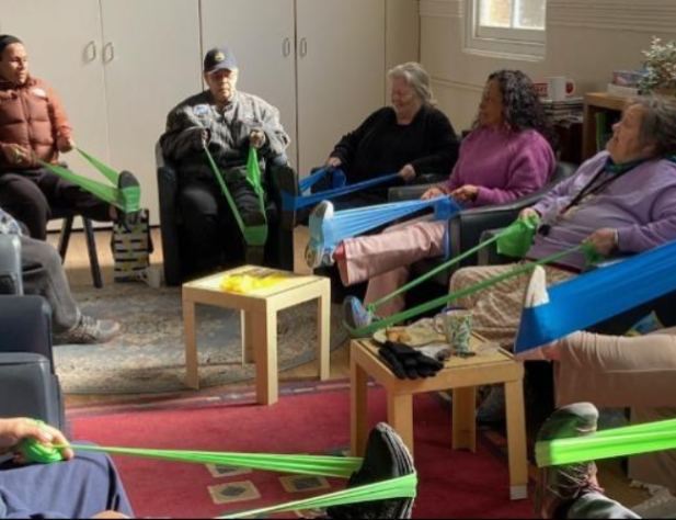 Nine people sitting in chairs in a circle, each using blue and green stretch bands, pulling against the arches of their feet