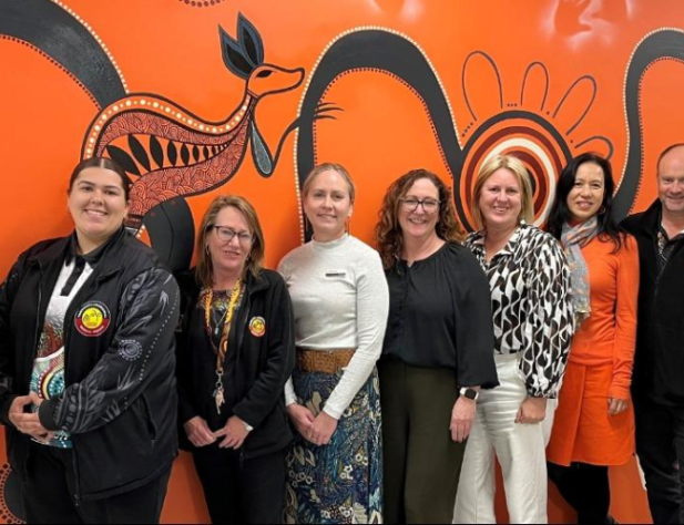 Nine people standing in a line in front of an orange Aboriginal artwork wall