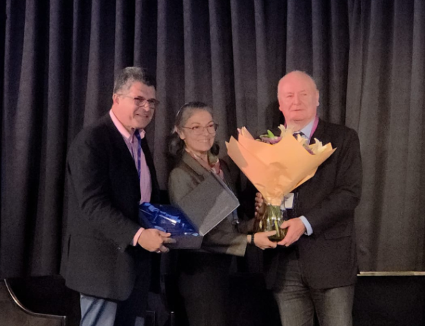 Man, woman and man standing on a stage with a black back drop and Optometry NSW/ACT sign, holding an award and flowers